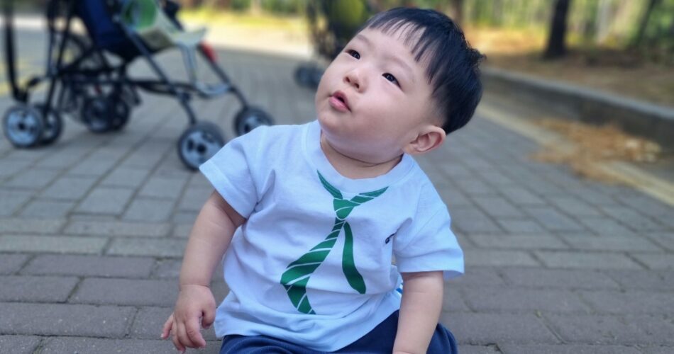 하얀색 셔츠와 파란 바지를 입고 녹색 타이 디자인이 있는 지호가 공원 포장도로에 앉아 하늘을 올려다보고 있다. 뒤편에는 유모차가 흐릿하게 보인다.