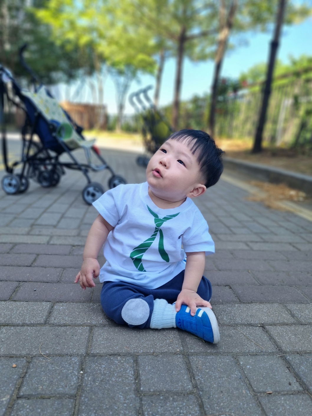 하얀색 셔츠와 파란 바지를 입고 녹색 타이 디자인이 있는 지호가 공원 포장도로에 앉아 하늘을 올려다보고 있다. 뒤편에는 유모차가 흐릿하게 보인다.