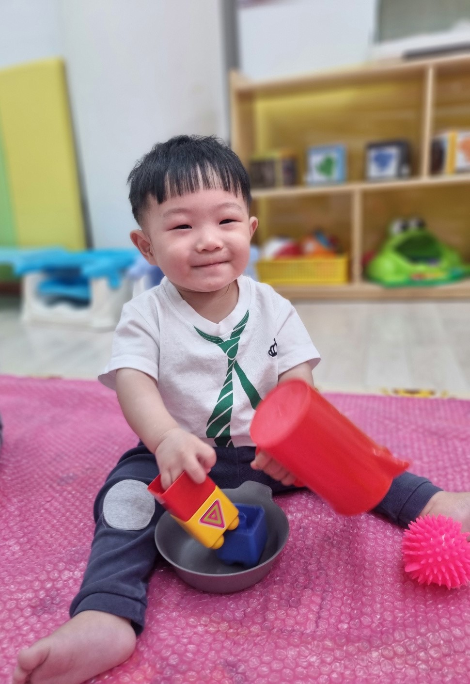 흰색 티셔츠와 초록색 넥타이 무늬를 입은 아기 지호가 분홍색 매트 위에 앉아 빨간색 컵과 노란색 블록, 금속 그릇을 가지고 놀며 활짝 웃고 있는 모습.