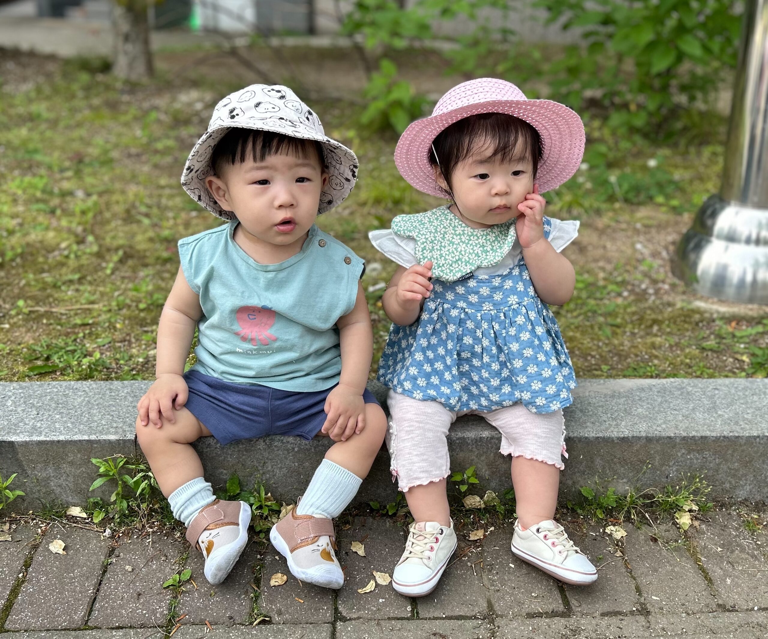 지호가 민트색 옷과 모자를 쓰고 다른 아기 친구와 함께 공원 벤치에 나란히 앉아있다. 친구는 분홍색 모자와 꽃무늬 원피스를 입고 있다.