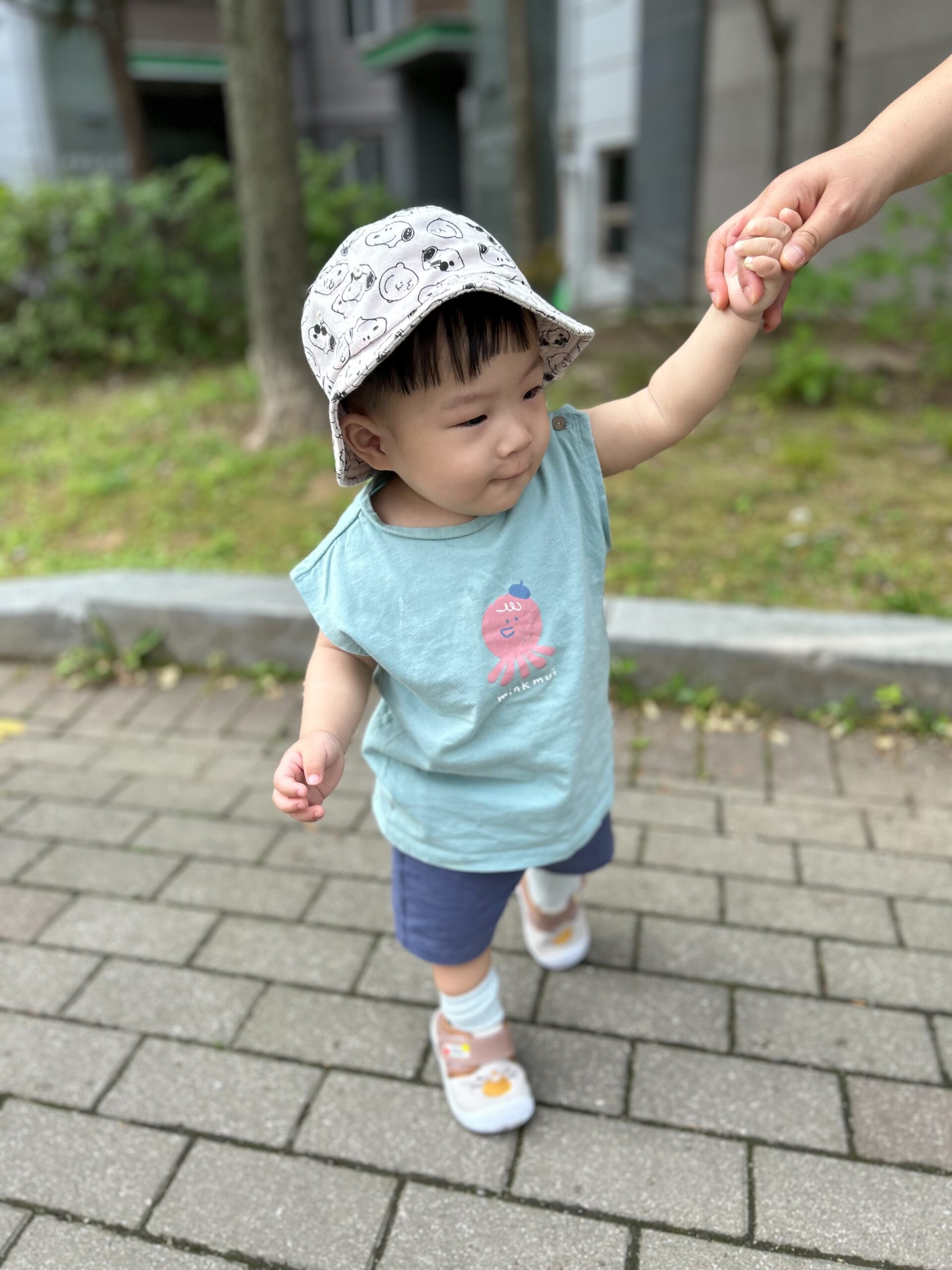 민트색 민소매 티셔츠와 파란색 반바지, 귀여운 모자를 쓴 지호가 어른의 손을 꼭 잡고 공원 길을 걷고 있다. 초록색 배경이 싱그럽다.