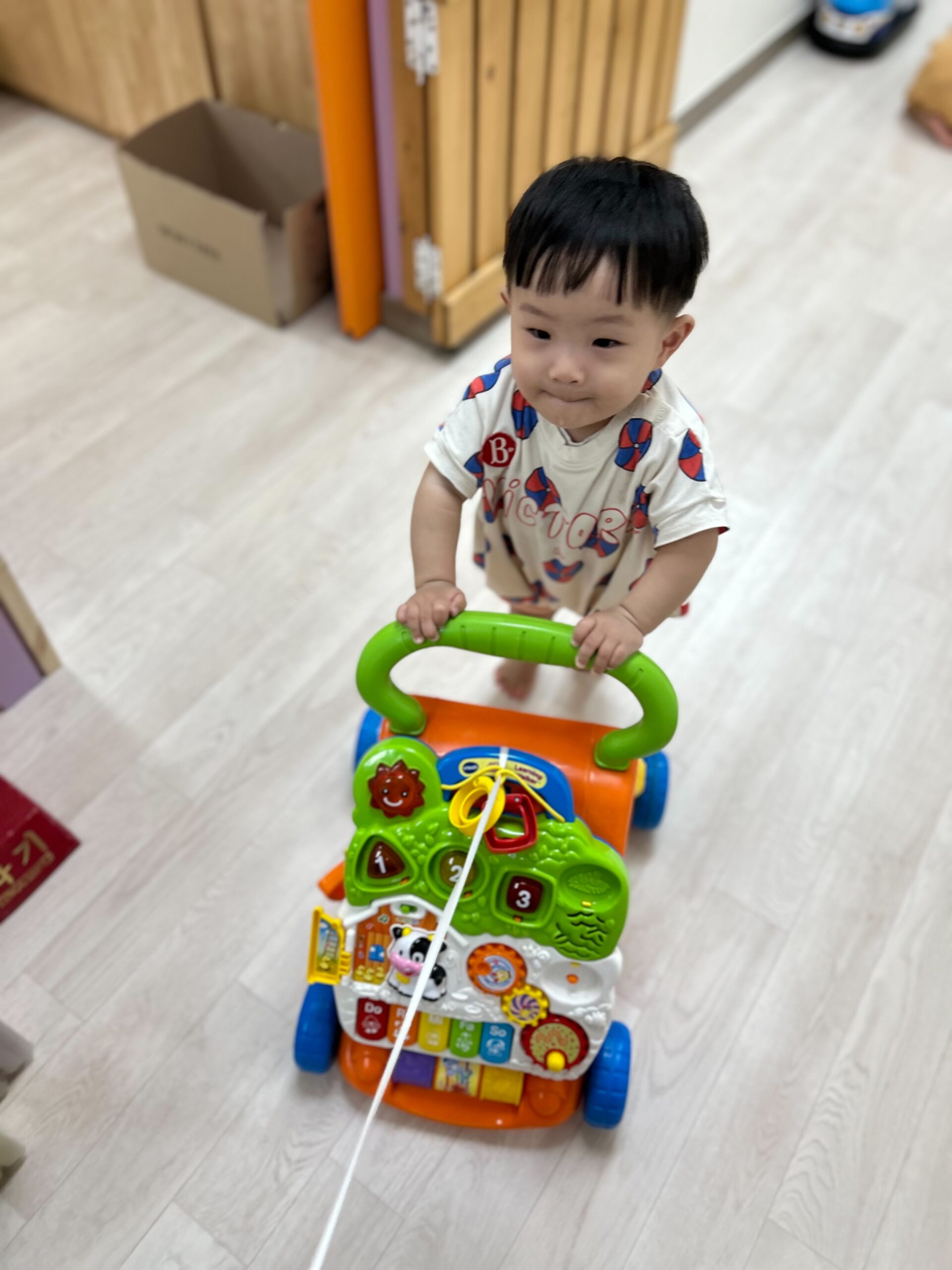 실내에서 알록달록한 걸음마 보조기를 잡고 서 있는 지호의 모습. 아래를 내려다보며 집중하고 있다.