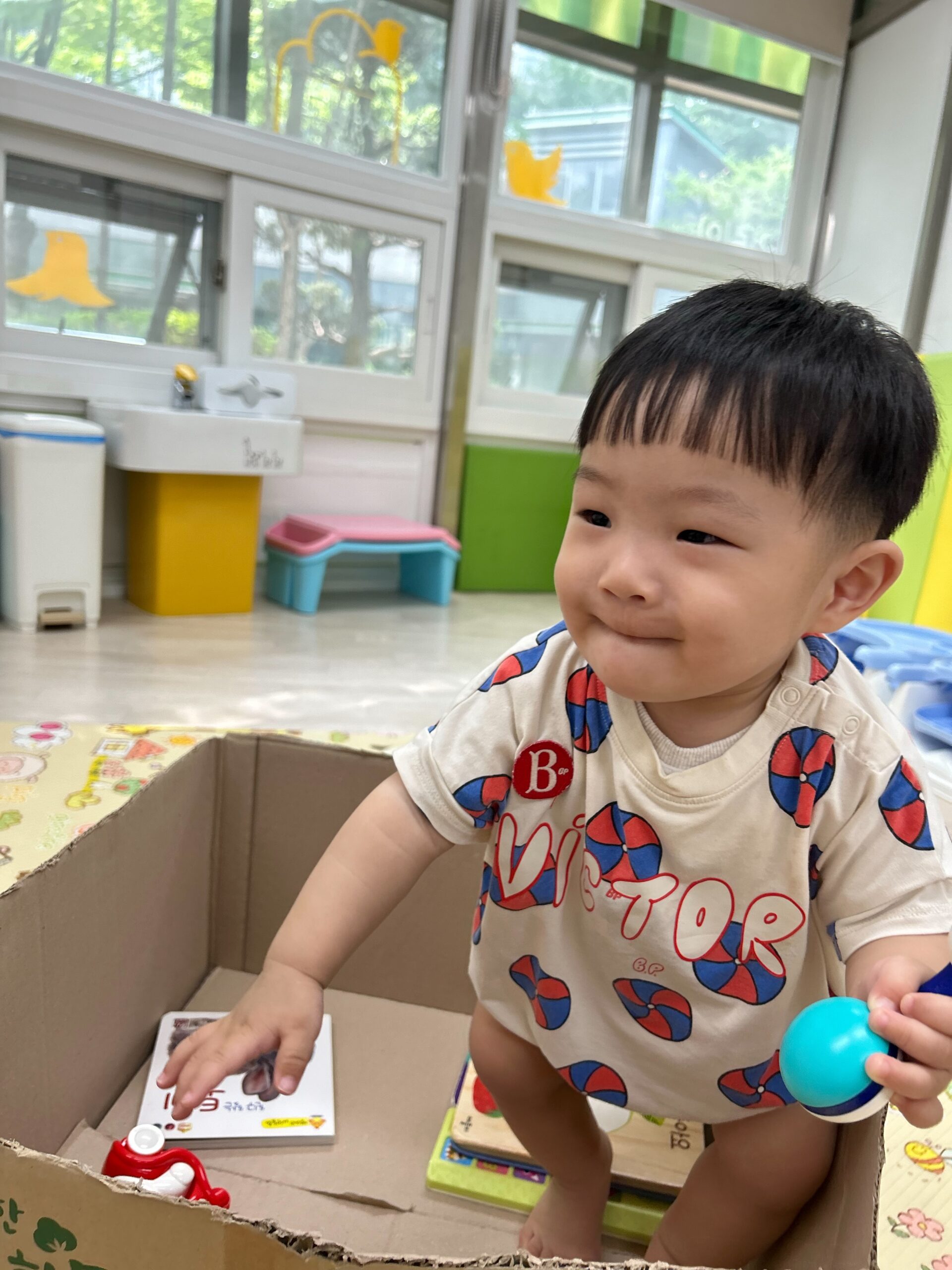 커다란 골판지 상자 안에 서서 작은 파란색 장난감을 쥐고 환하게 웃고 있는 지호의 상반신 사진. 흰색 바탕에 빨간색과 파란색 무늬가 있는 롬퍼를 입고 있다. 배경에는 창문 밖 푸른 나무와 실내 놀이 공간의 가구들이 보인다.