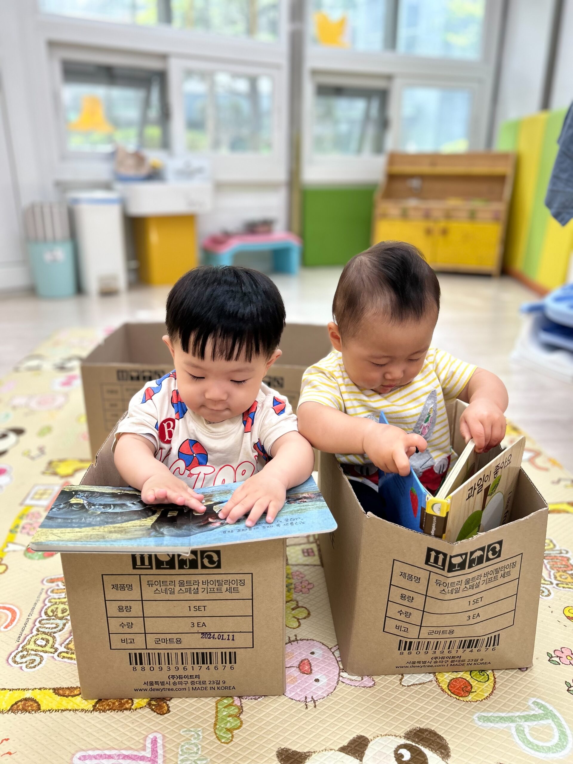 두 명의 아기가 각각 골판지 상자 안에 앉아 책을 보고 있는 모습. 지호는 왼쪽에 앉아 파란색 표지의 책을 보고 있고, 오른쪽 아기는 노란색 줄무늬 옷을 입고 다른 책을 보고 있다. 두 아기 모두 책에 집중하고 있으며, 배경은 밝은 실내 놀이 공간이다.