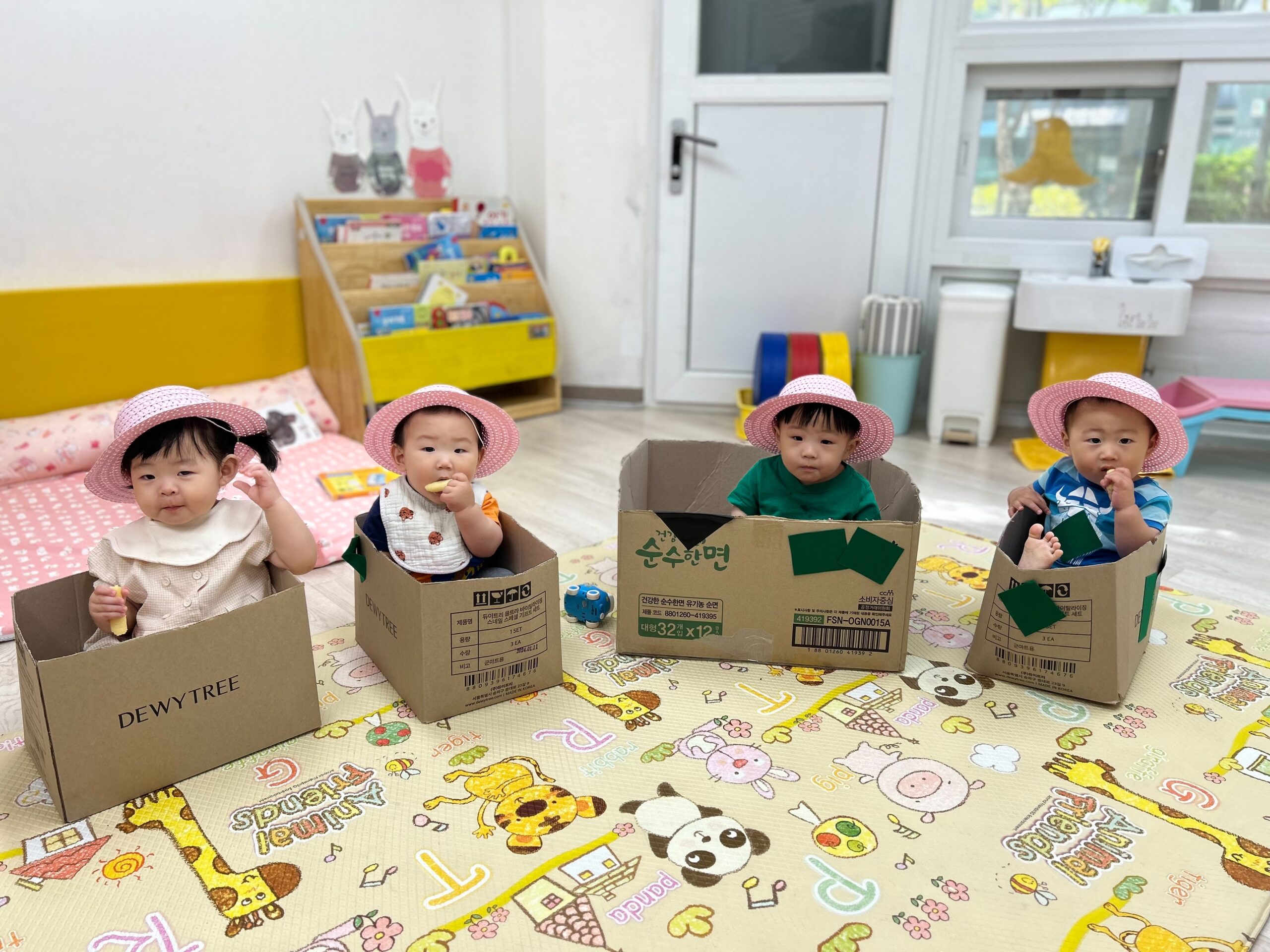 어린이집 실내에서 네 명의 아기들이 분홍색 밀짚모자를 쓰고 각자 다른 디자인의 골판지 상자 안에 앉아 카메라를 바라보고 있다. 지호는 왼쪽에서 세 번째에 위치한다.