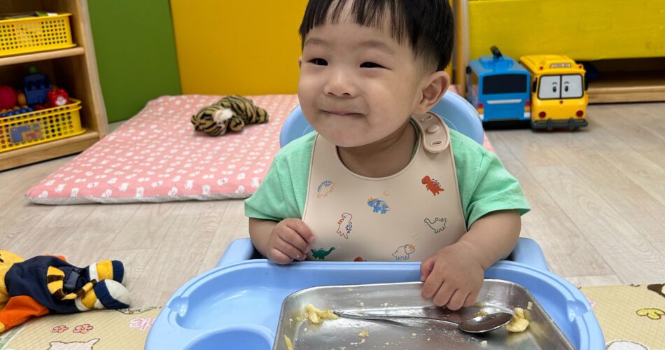 초록색 티셔츠와 공룡 그림 턱받이를 한 지호가 유아용 식탁의자에 앉아 접시 위 바나나 조각을 다 먹고 만족스러운 미소를 짓고 있다. 뒤쪽으로는 낮은 책장과 놀이방 매트가 보인다.