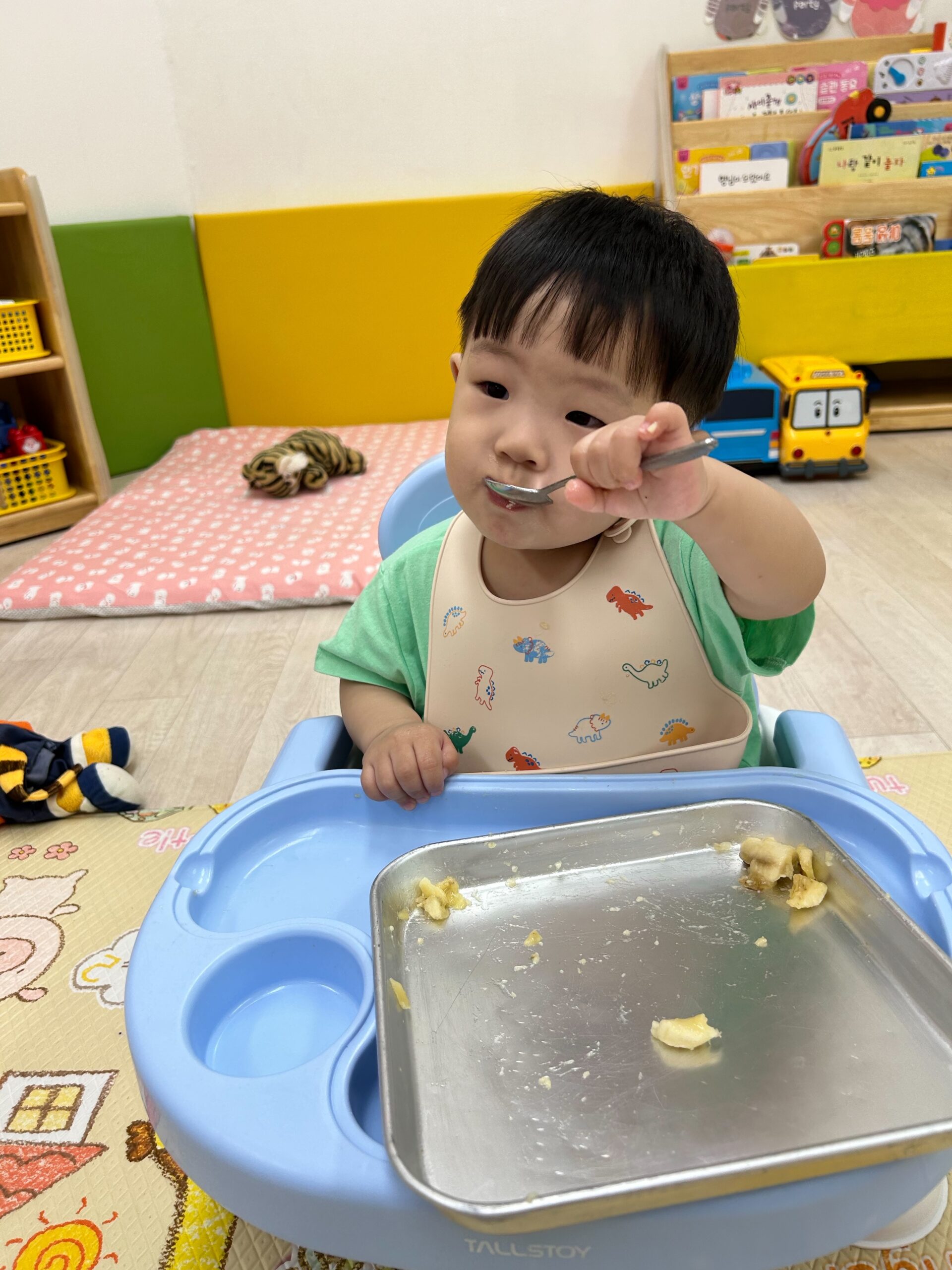 연두색 티셔츠와 공룡 무늬 턱받이를 한 지호가 파란색 유아용 식탁의자에 앉아 숟가락으로 음식을 떠먹고 있다. 앞에는 음식이 담긴 은색 트레이가 놓여 있다.