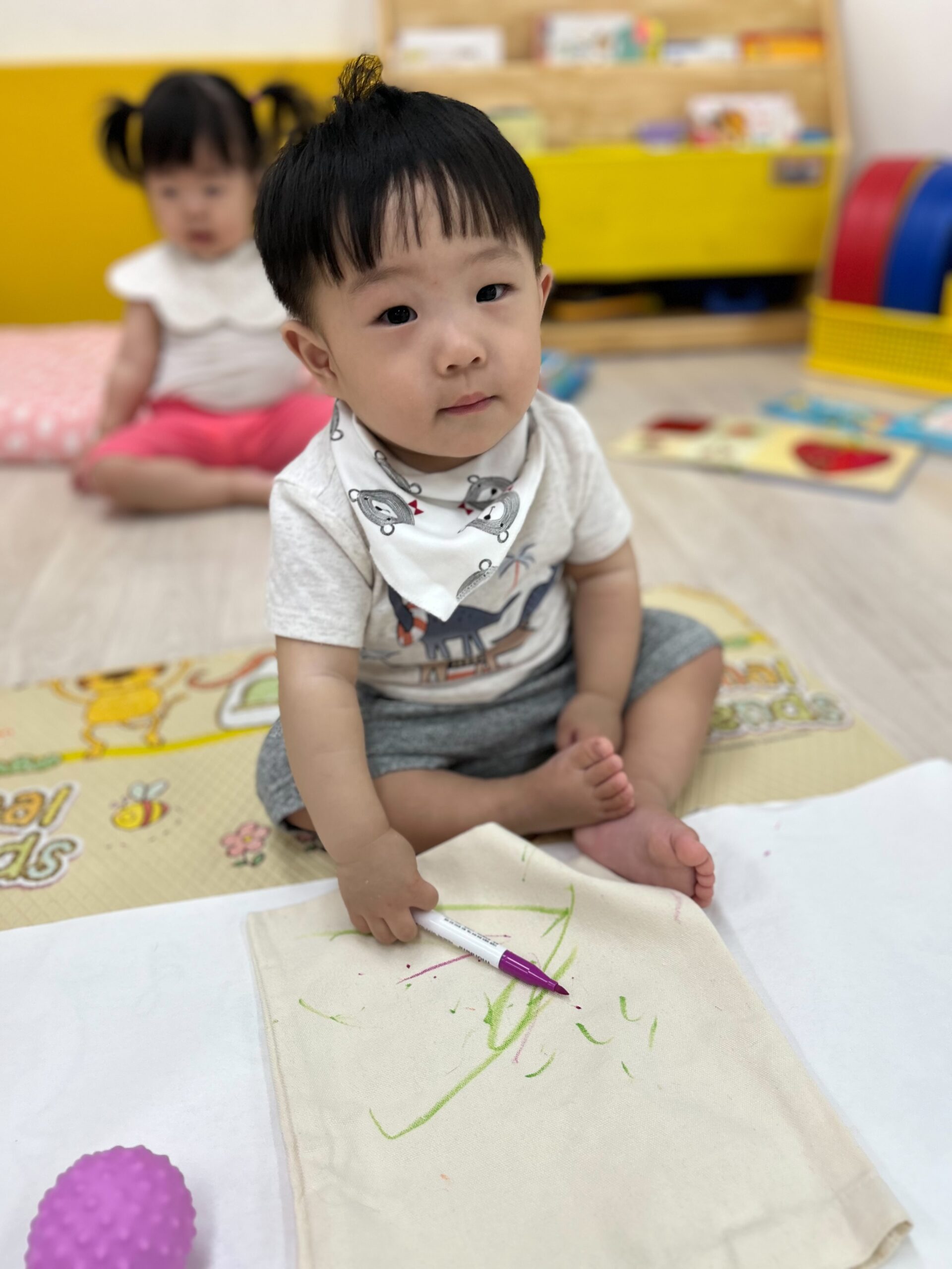 실내 바닥에 앉아 회색 티셔츠와 턱받이를 한 지호가 보라색 마커로 하얀 천에 그림을 그리고 있다. 지호는 카메라를 바라보고 있으며, 뒤편에는 다른 아이가 흐릿하게 앉아있다.