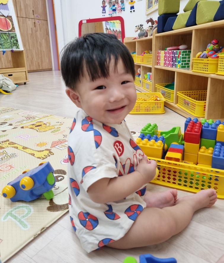 귀여운 패턴의 실내복을 입은 지호가 알록달록한 블록들이 담긴 노란색 바구니 옆에 앉아 카메라를 보며 환하게 웃고 있다. 바닥에는 동물 그림 매트가 깔려 있다.