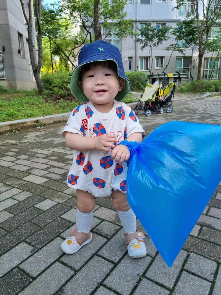 파란색 버킷햇을 쓰고 흰색 패턴의 옷을 입은 지호가 아파트 단지 보도블록 위에서 파란색 비닐봉투를 손목에 걸고 환하게 웃으며 서 있는 모습. 뒤로는 유모차 두 대가 보인다.