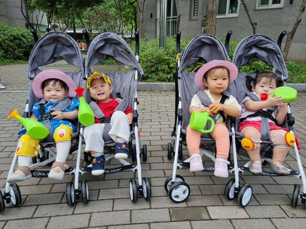 네 명의 아기들이 회색 유모차에 나란히 앉아 야외에서 카메라를 보고 있다. 왼쪽부터 파란색 옷의 지호는 노란색 모자를 쓰고 환하게 웃고 있으며, 다른 아기들도 각자 모자와 장난감을 들고 있다.