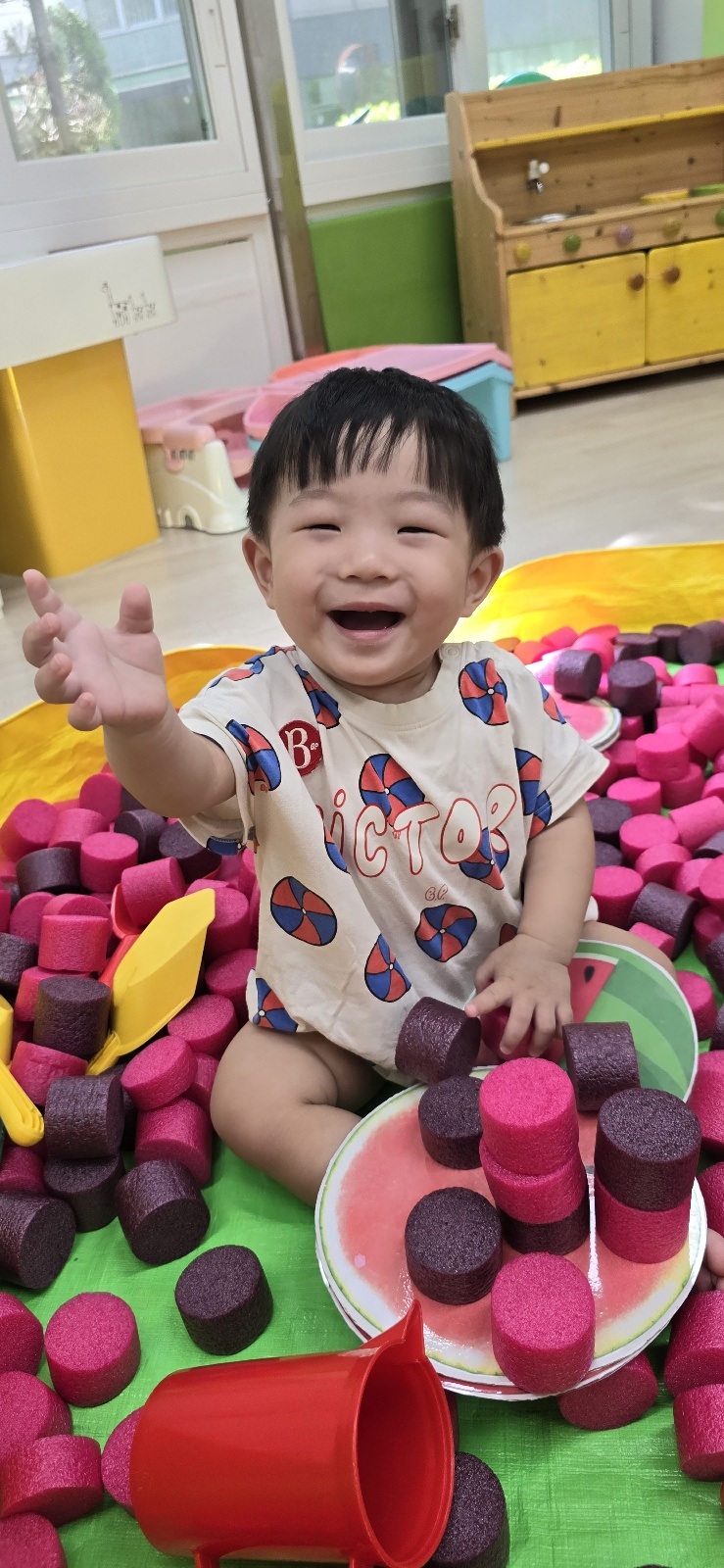 분홍색과 자주색의 원통형 스펀지 블록으로 가득 찬 공간에 앉아 해맑게 웃으며 손을 내밀고 있는 지호의 모습. 노란색 접시와 삽도 보인다.