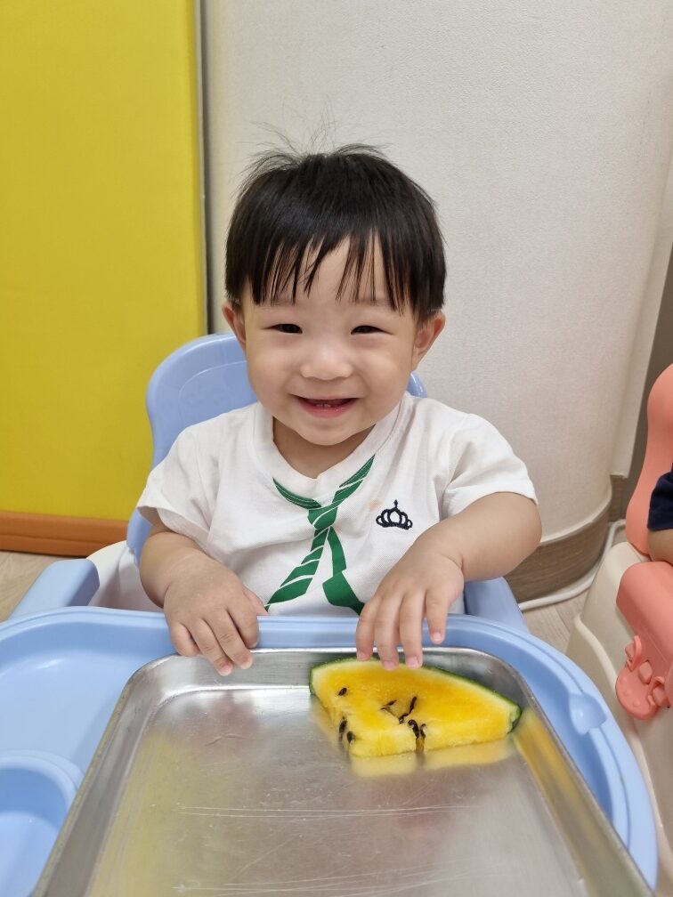 실내에서 파란색 아기 식탁 의자에 앉아 밝게 웃고 있는 지호. 흰색 티셔츠에 초록색 넥타이 무늬가 그려져 있으며, 앞의 금속 트레이 위에는 씨가 있는 노란색 수박 한 조각이 놓여 있다.