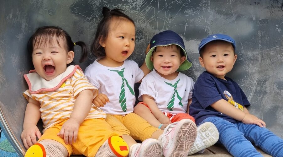 야외 놀이터의 미끄럼틀 위에서 네 명의 아기들이 나란히 앉아 활짝 웃고 있다. 왼쪽 첫 번째 아기 지호는 노란색과 흰색 줄무늬 상의를 입고 노란색 무릎 보호대를 착용한 채 입을 크게 벌리고 웃고 있으며, 그 옆의 아기는 초록색 넥타이 무늬 흰색 티셔츠를 입고 입을 벌린 채 무언가를 말하는 듯하다. 오른쪽 두 명의 아기들도 밝게 미소 짓고 있다.