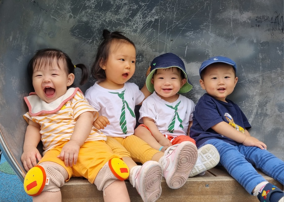 야외 놀이터의 미끄럼틀 위에서 네 명의 아기들이 나란히 앉아 활짝 웃고 있다. 왼쪽 첫 번째 아기 지호는 노란색과 흰색 줄무늬 상의를 입고 노란색 무릎 보호대를 착용한 채 입을 크게 벌리고 웃고 있으며, 그 옆의 아기는 초록색 넥타이 무늬 흰색 티셔츠를 입고 입을 벌린 채 무언가를 말하는 듯하다. 오른쪽 두 명의 아기들도 밝게 미소 짓고 있다.