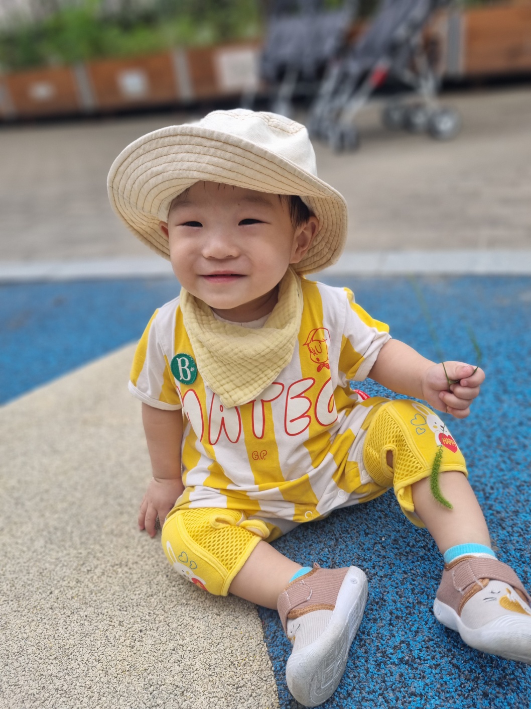 야외 놀이터 바닥에 앉아 카메라를 보고 환하게 웃는 지호의 모습. 베이지색 벙거지 모자와 노란색 줄무늬 롬퍼를 입고 있으며, 한 손에는 작은 나뭇잎 같은 것을 들고 있다. 배경에는 유모차가 흐릿하게 보인다.