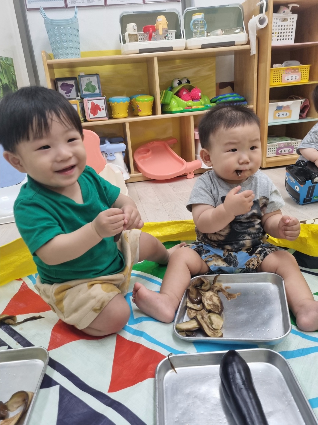 초록색 티셔츠를 입은 지호가 바닥에 앉아 친구와 함께 금속 쟁반에 담긴 삶은 가지 조각들을 만지고 있다. 지호는 활짝 웃고 있으며, 친구는 가지를 입에 대고 있다.
