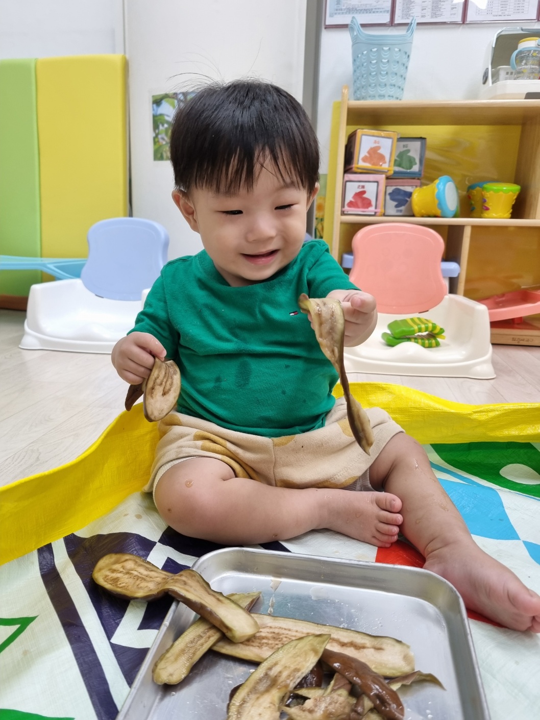 초록색 티셔츠를 입은 지호가 바닥에 앉아 삶은 가지 조각들을 양손에 들고 진지한 표정으로 내려다보고 있다. 바닥에는 다채로운 패턴의 매트가 깔려 있다.