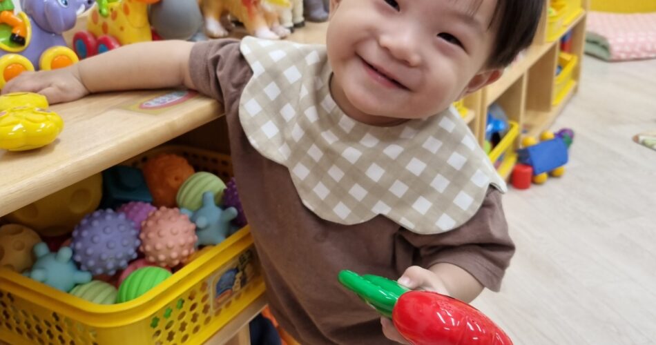밝은 갈색 옷과 체크무늬 턱받이를 한 지호가 장난감 선반에 기대어 서서 환하게 웃으며 빨간색 당근 장난감을 들고 있다. 주변에는 다양한 색깔의 장난감들이 보인다.