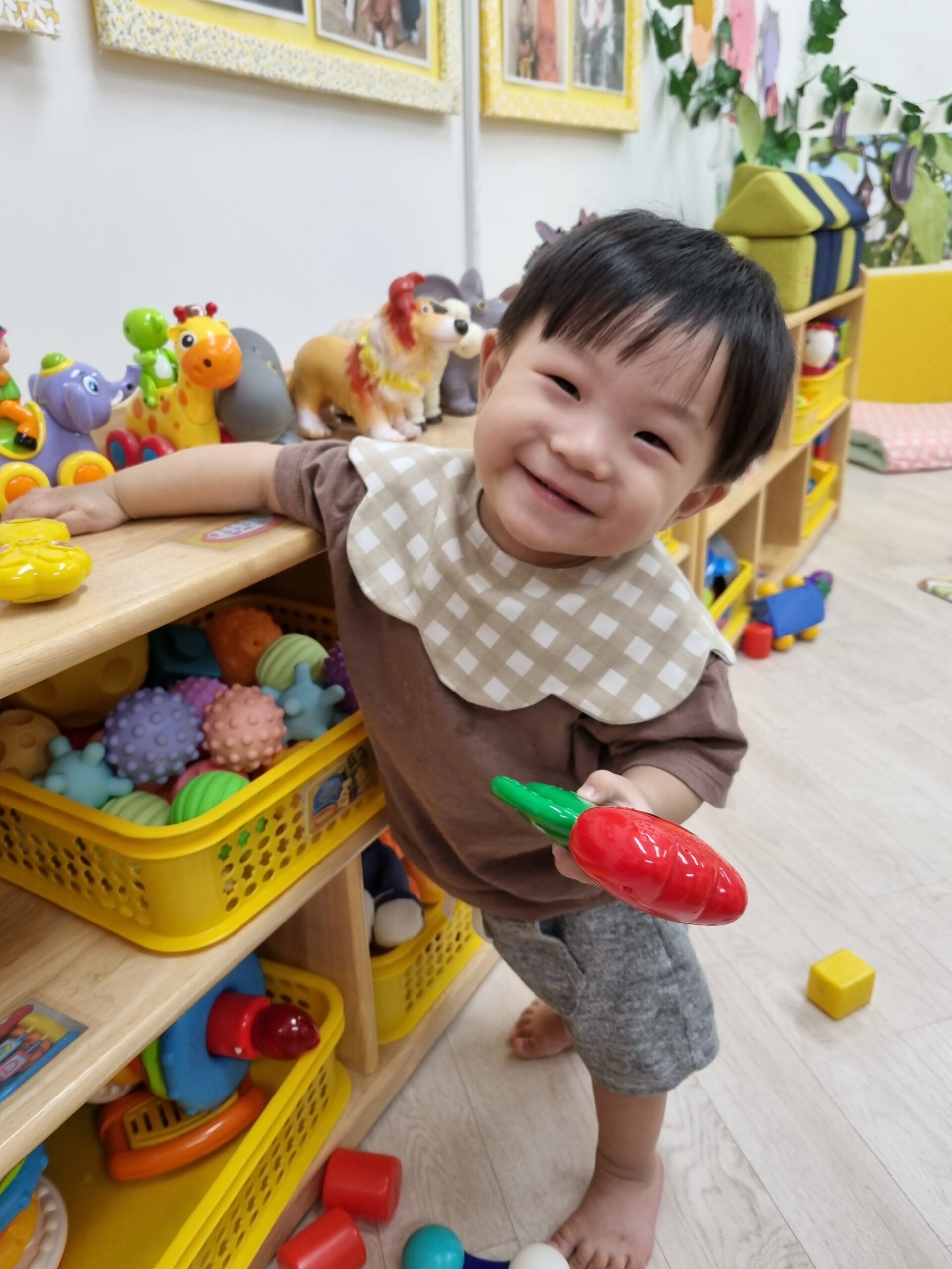 밝은 갈색 옷과 체크무늬 턱받이를 한 지호가 장난감 선반에 기대어 서서 환하게 웃으며 빨간색 당근 장난감을 들고 있다. 주변에는 다양한 색깔의 장난감들이 보인다.
