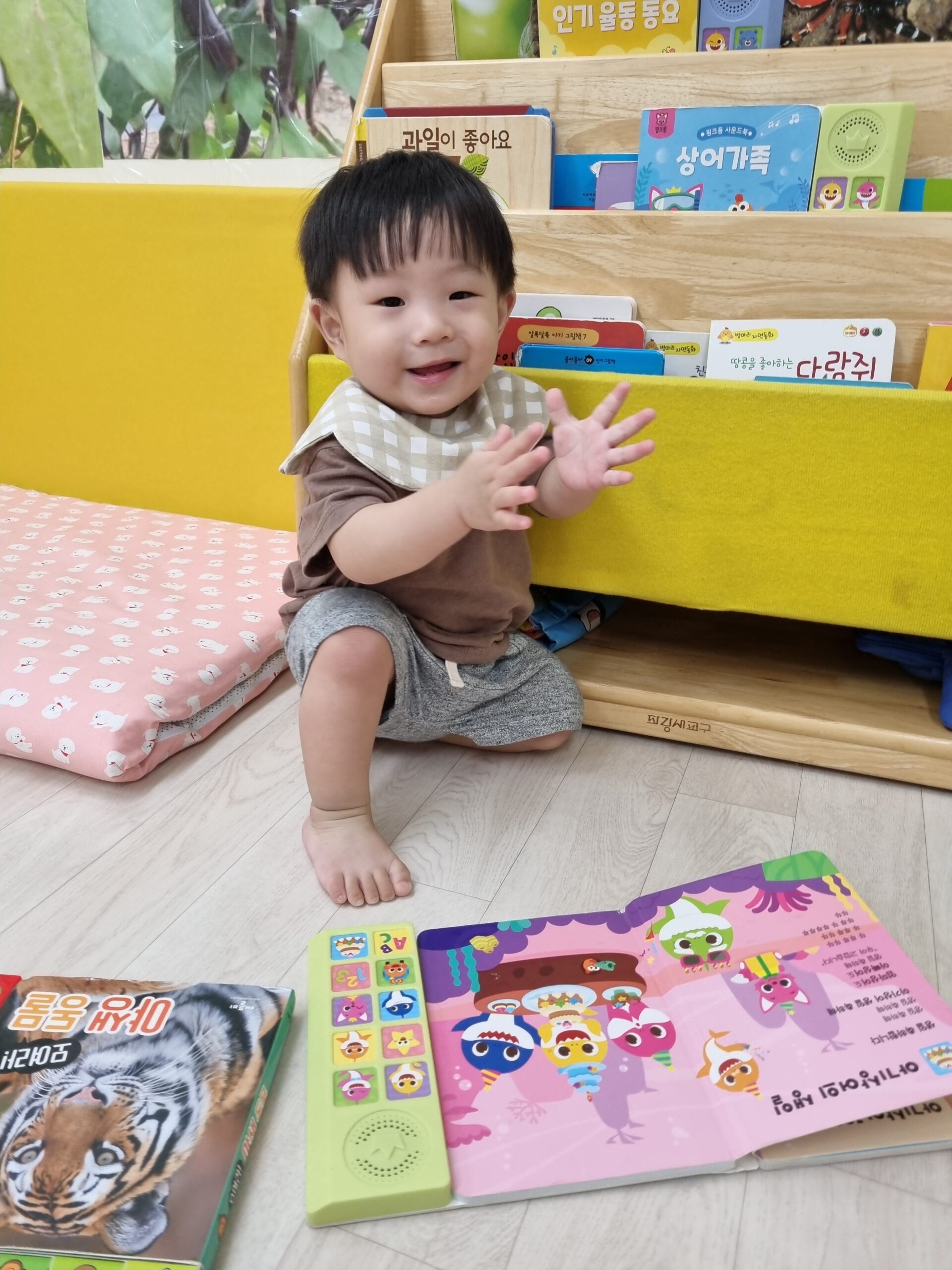 노란색 책장 앞에 앉아 갈색 상의와 회색 하의, 체크무늬 턱받이를 한 지호가 활짝 웃으며 양손으로 박수를 치고 있는 모습. 앞에는 소리 나는 그림책과 다른 책이 놓여 있다.