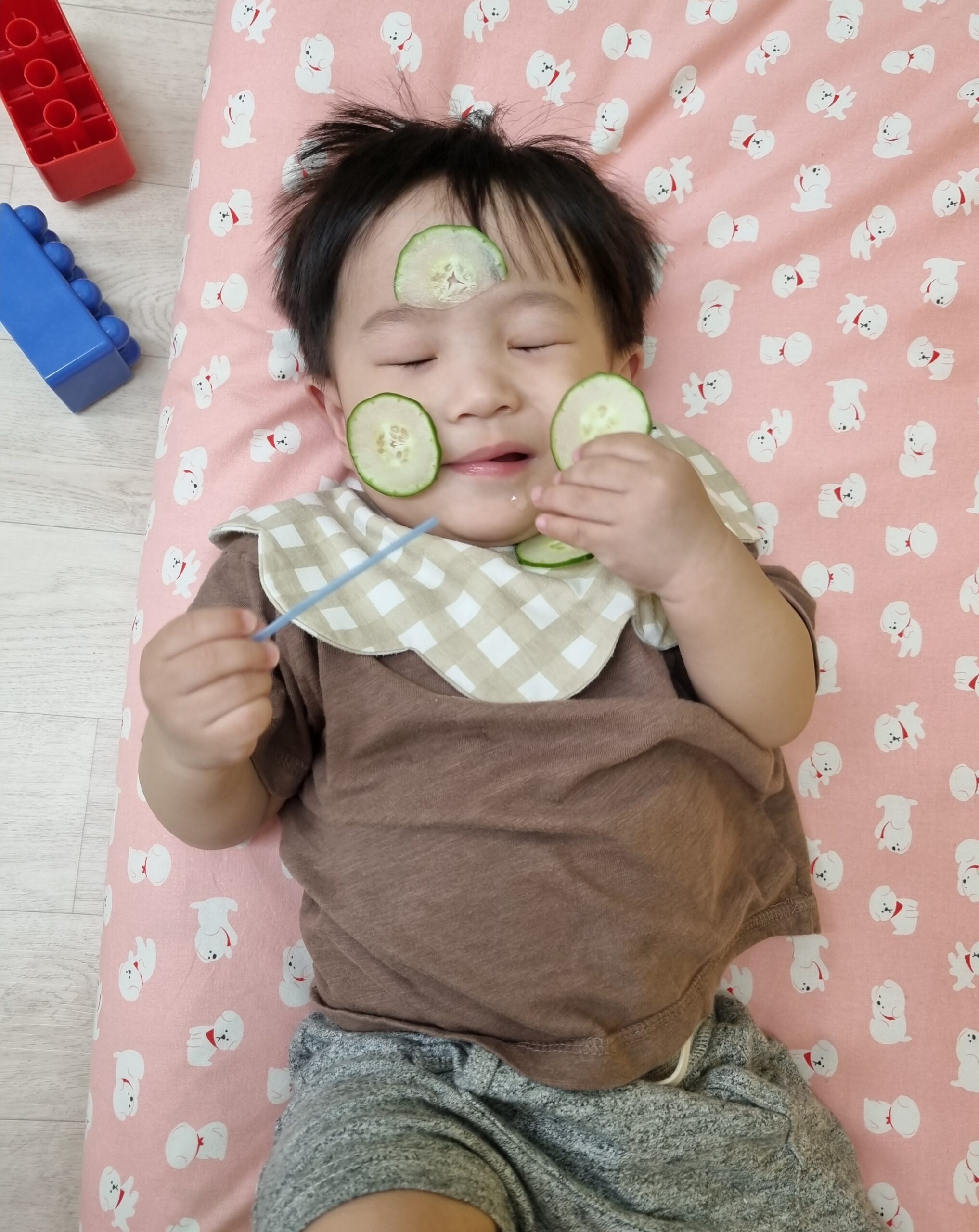 분홍색 패턴 매트에 누워있는 지호의 얼굴에 오이 슬라이스가 올려져 있고, 지호는 눈을 감은 채 편안한 표정으로 오른손에 긴 막대를, 왼손에 오이 한 조각을 들고 있다.