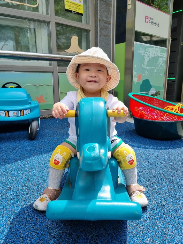 화창한 야외에서 파란색 말 모양의 흔들 목마에 앉아 정면을 바라보며 환하게 미소 짓는 지호. 베이지색 벙거지 모자와 노란색 줄무늬 롬퍼, 노란색 무릎 보호대를 착용하고 있다. 뒤편에는 건물과 다른 놀이 시설들이 보인다.