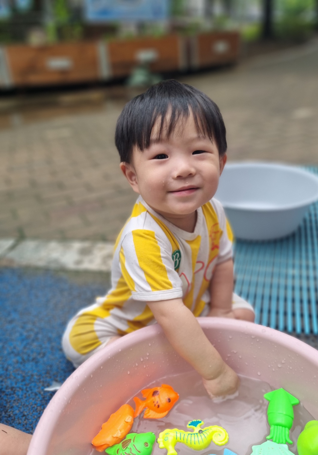 노란색과 흰색 줄무늬 옷을 입은 아기가 야외 파란색 매트 위에 앉아 분홍색 대야에 담긴 물에 손을 넣고 물고기 모양 장난감들과 함께 즐거운 물놀이를 하고 있다. 행복하게 카메라를 바라보며 웃고 있다.