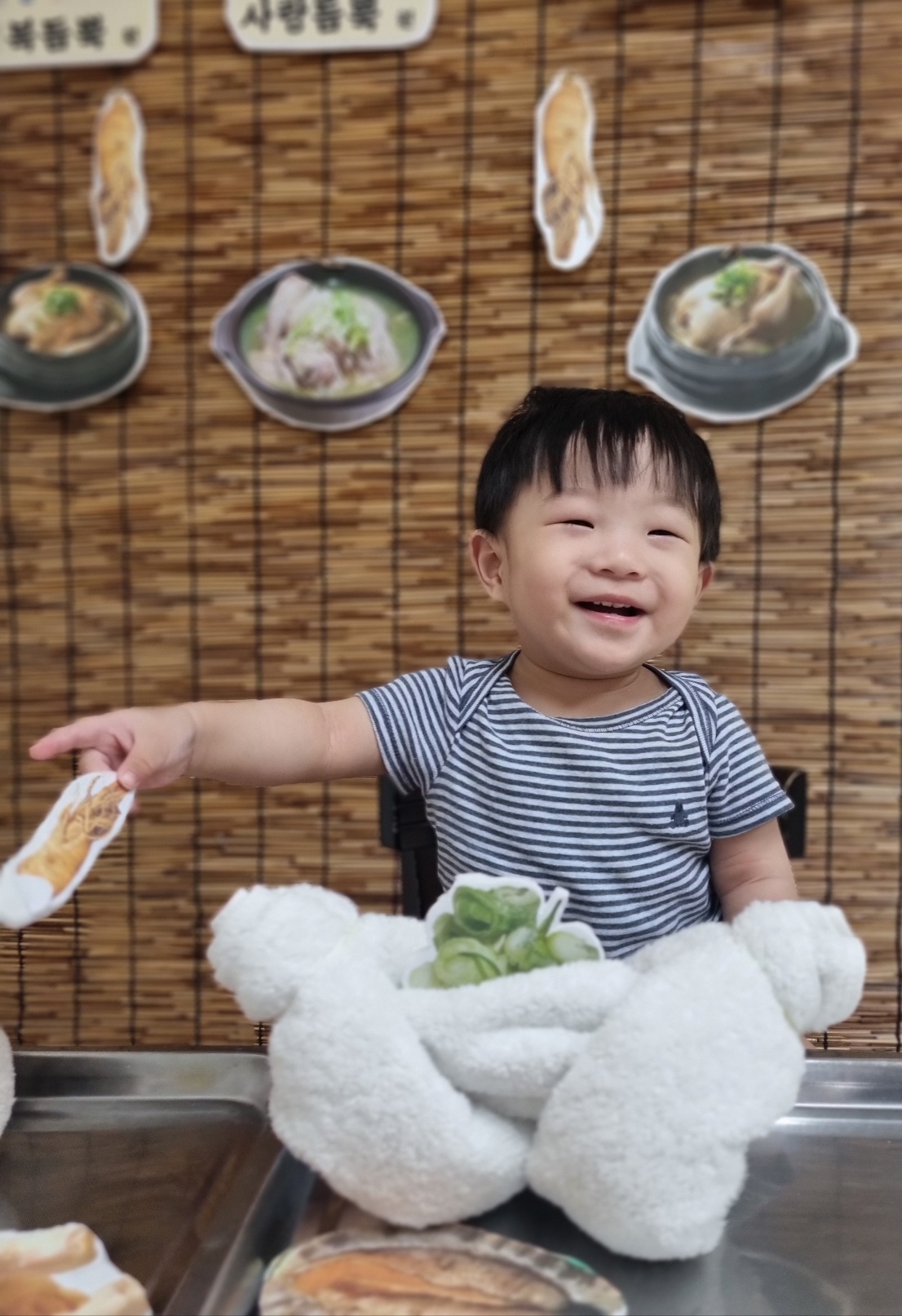 줄무늬 티셔츠를 입은 아기가 대나무 배경 앞에서 환하게 웃으며 손가락으로 무언가를 가리키고 있다. 앞에는 부드러운 흰색 수건으로 만든 장난감이 놓여있고, 배경에는 삼계탕과 전복죽 사진이 걸려 있다.