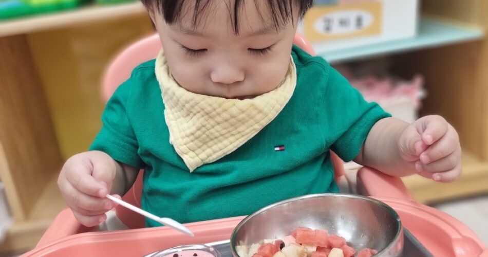 초록색 티셔츠와 노란색 턱받이를 한 아기가 아기 의자에 앉아 작은 숟가락으로 그릇에 담긴 수박 조각을 조심스럽게 떠서 먹으려 집중하고 있다. 주변에는 다른 음식 그릇들도 보인다.