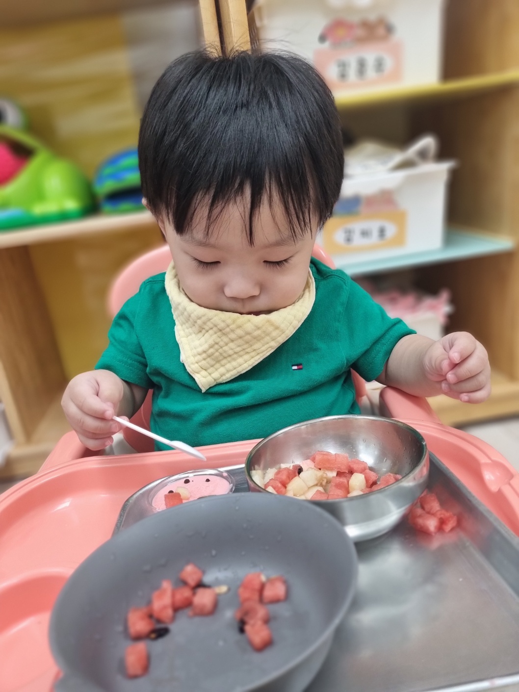 초록색 티셔츠와 노란색 턱받이를 한 아기가 아기 의자에 앉아 작은 숟가락으로 그릇에 담긴 수박 조각을 조심스럽게 떠서 먹으려 집중하고 있다. 주변에는 다른 음식 그릇들도 보인다.