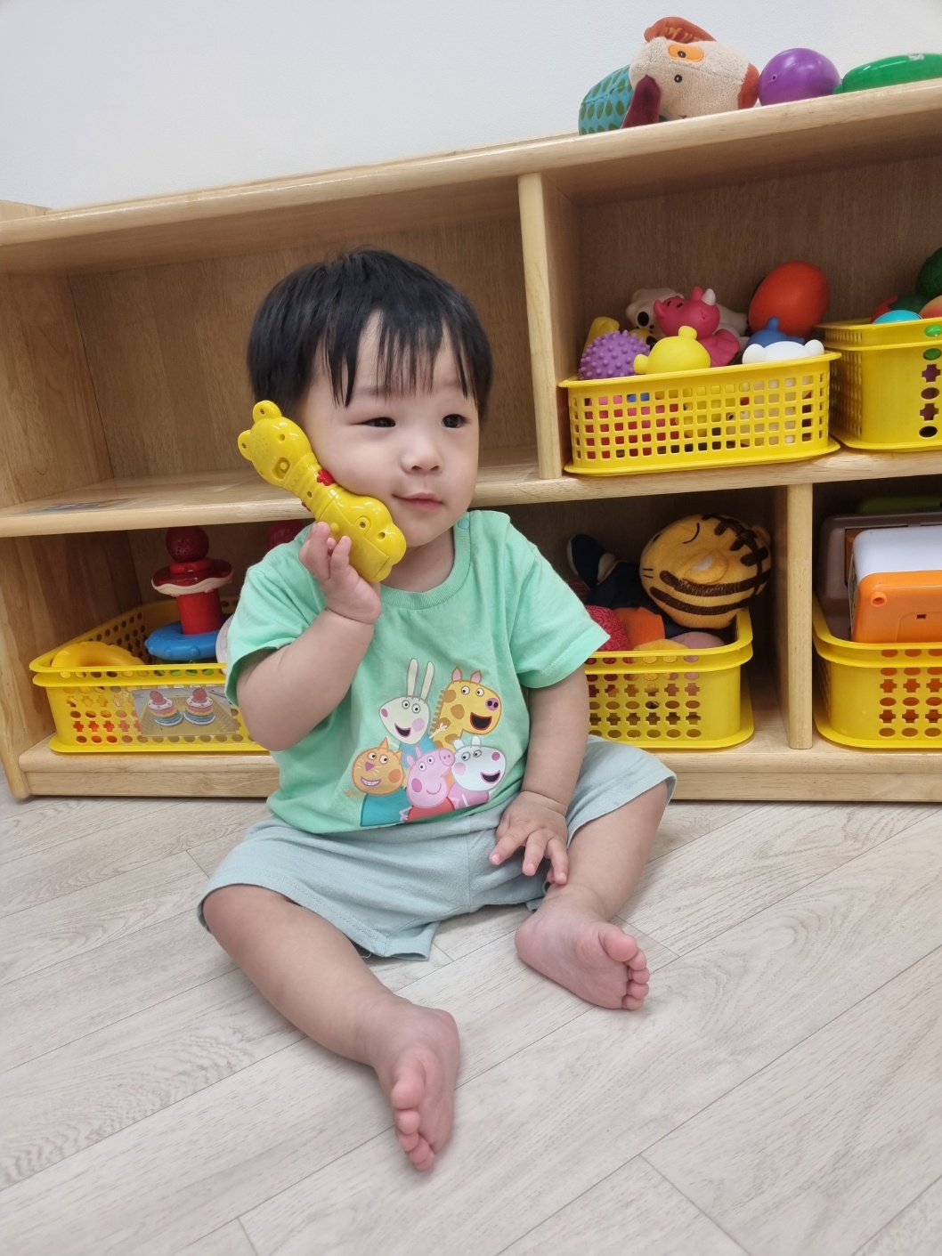 어린이집 실내 놀이방에서 아기 지호가 초록색 티셔츠를 입고 바닥에 앉아 노란색 장난감 전화기를 귀에 대고 사뭇 진지한 표정으로 어딘가에 이야기하는 듯한 포즈를 취하고 있다. 뒤편에는 다양한 장난감들이 놓여 있다.