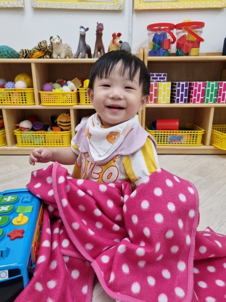 어린이집 실내 놀이방에서 아기 지호가 분홍색 물방울 무늬 담요를 덮고 바닥에 앉아 환하게 웃으며 카메라를 바라보고 있다. 뒤편에는 장난감으로 가득 찬 노란색 바구니들이 진열되어 있다.