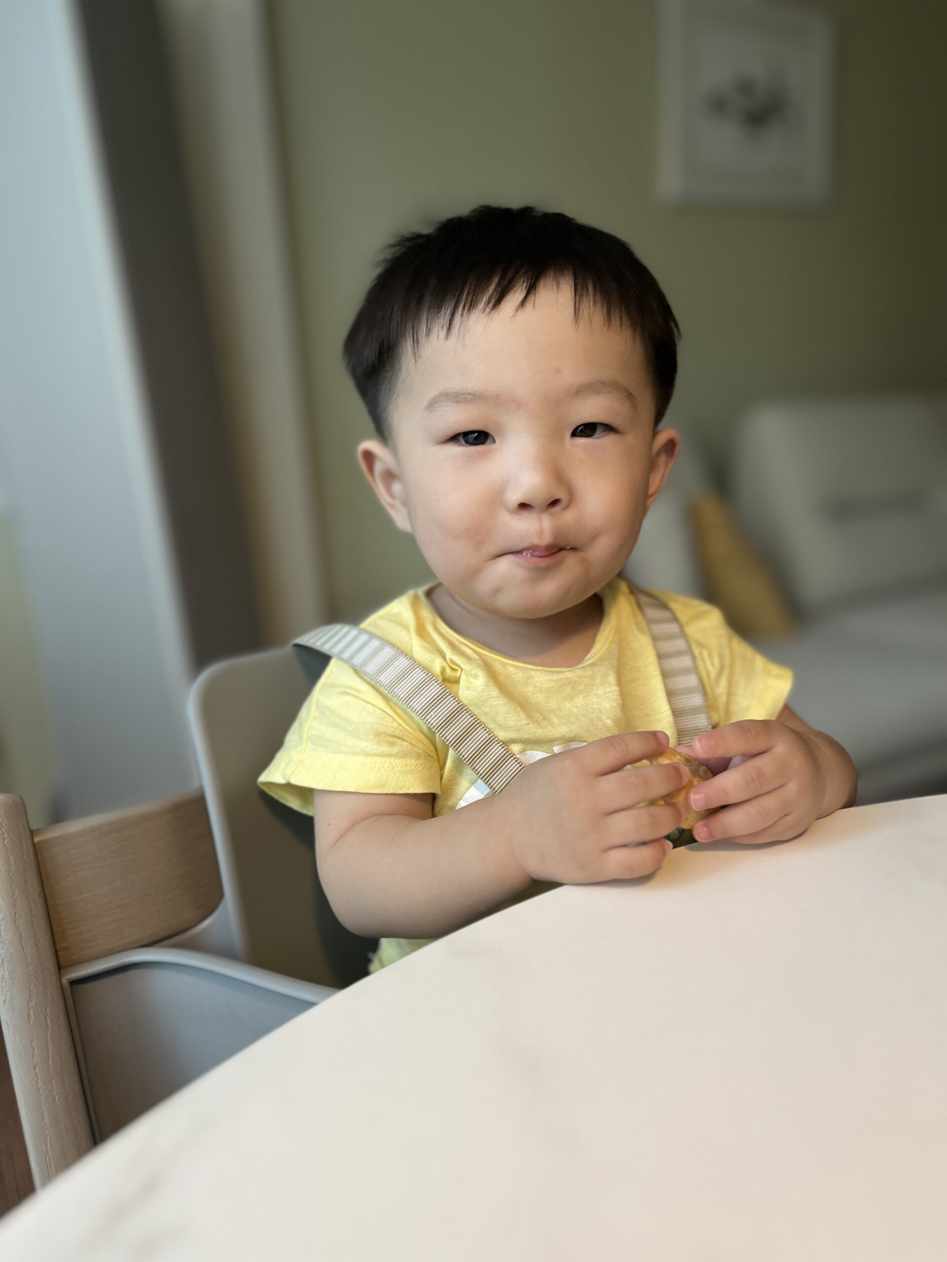 노란색 티셔츠와 멜빵을 입은 아기 지호가 하이체어에 앉아 흰색 식탁에 팔을 올리고 카메라를 보며 입술을 오므린 귀여운 표정을 짓고 있다. 배경은 가정집 거실로 보인다.