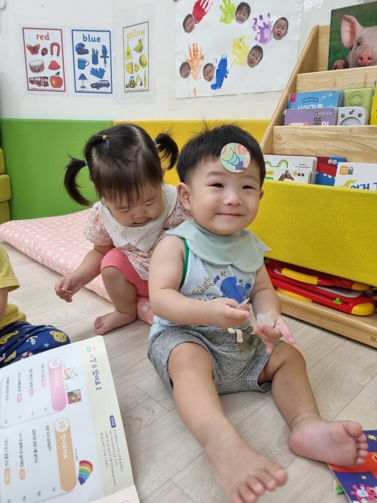 연두색 턱받이를 한 아기 지호가 바닥에 앉아 환하게 웃고 있으며, 이마에는 무지개 스티커가 붙어 있다. 옆에는 다른 여자아이가 고개를 숙여 책을 보고 있고, 뒤편에는 책장과 벽 그림이 보인다.