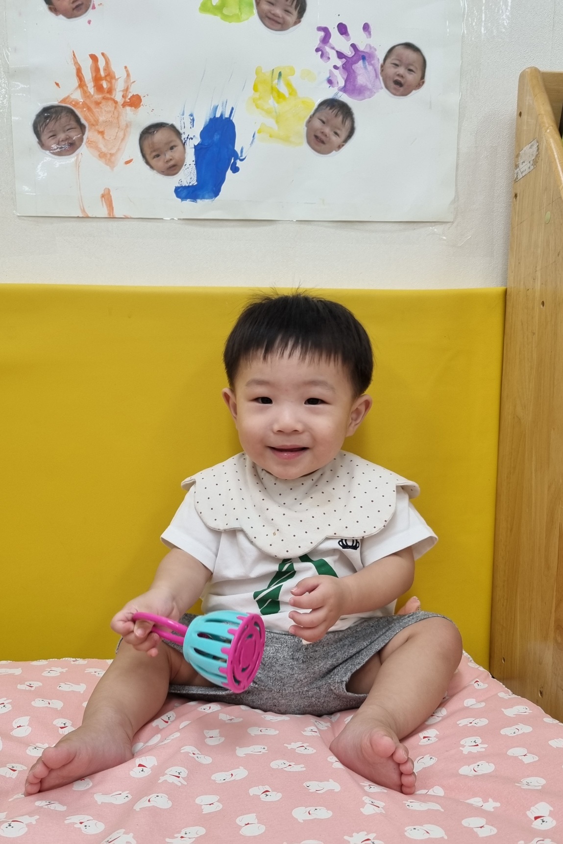 흰색 티셔츠와 물방울무늬 턱받이를 한 아기 지호가 노란색 벽과 분홍색 매트가 있는 어린이집 실내에서 앉아 카메라를 보고 환하게 웃고 있다. 손에는 알록달록한 장난감을 들고 있으며, 뒤 벽에는 아기 얼굴 사진과 손바닥 그림이 붙어 있다.