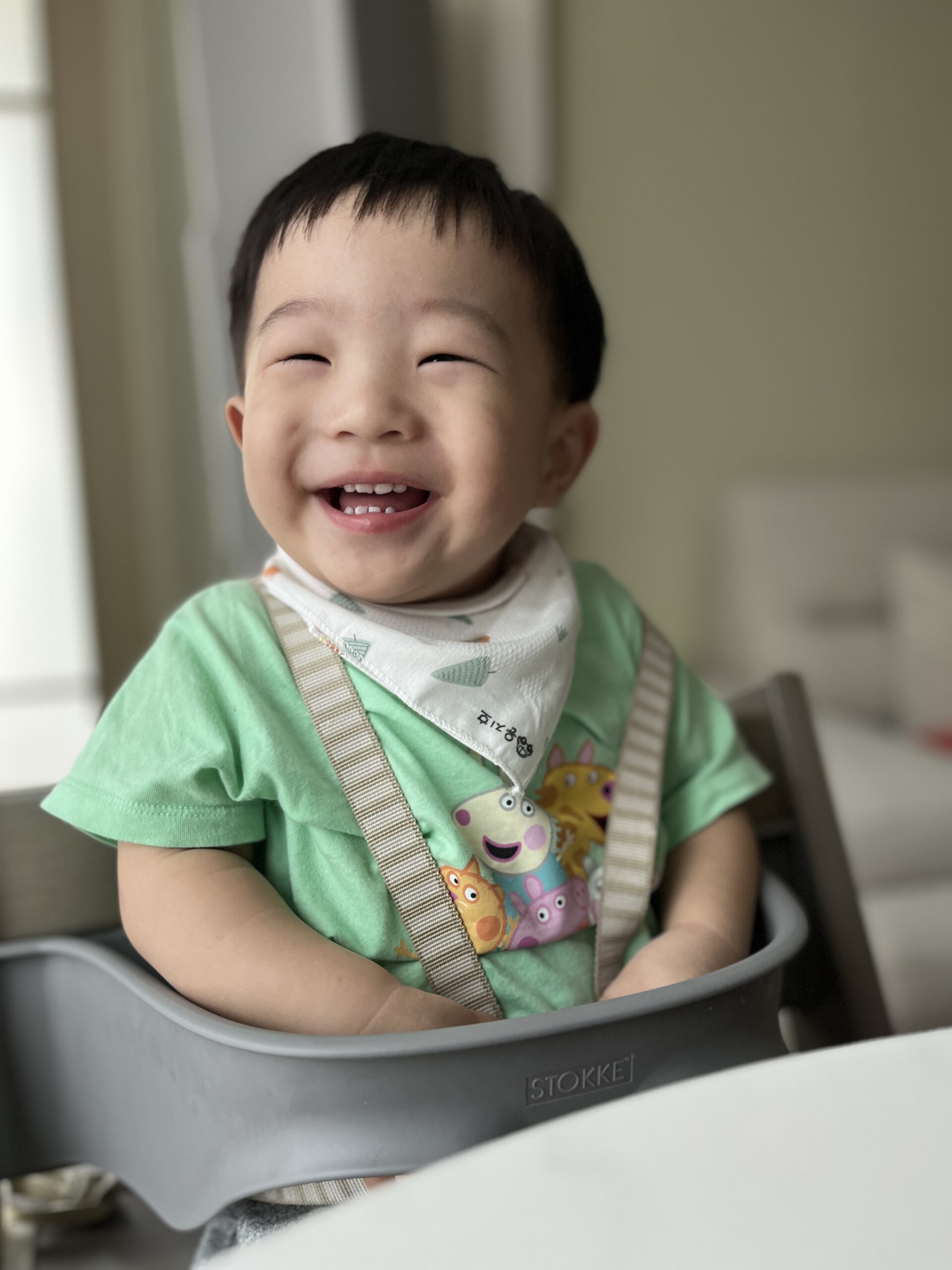 초록색 티셔츠와 귀여운 캐릭터 턱받이를 한 아기가 높은 아기 의자에 앉아 눈을 감고 입을 활짝 벌려 즐겁게 웃고 있다. 밝은 배경과 함께 지호의 행복한 순간이 포착되었다.