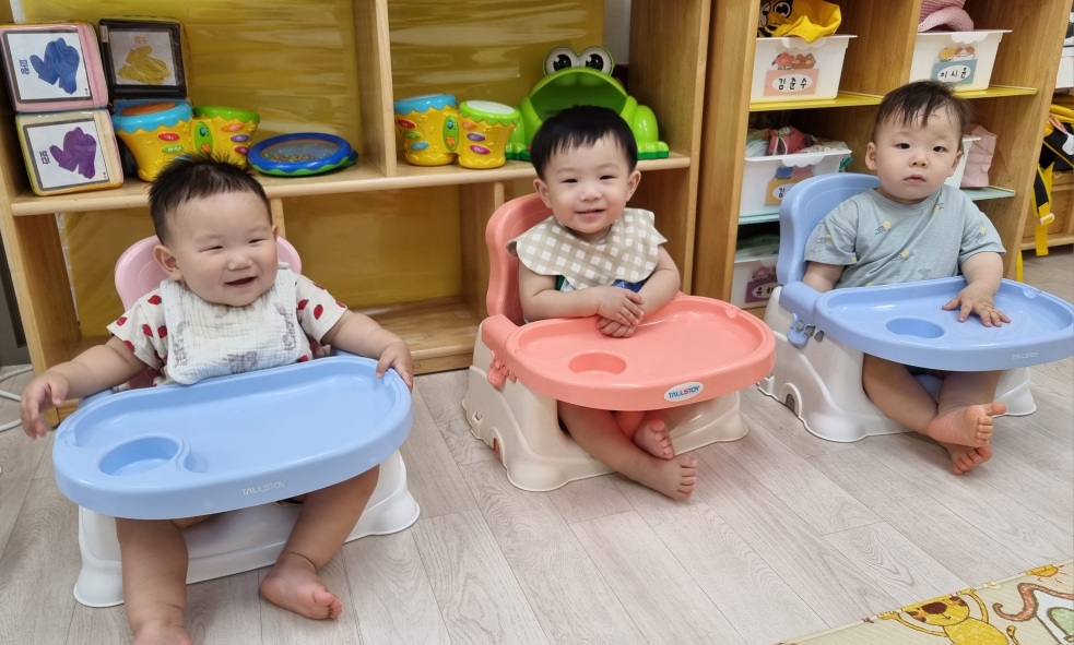 나무 바닥에 놓인 세 개의 아기 부스터 의자에 세 명의 아기가 나란히 앉아있다. 가운데 지호가 밝게 웃고 있으며, 양옆의 친구들도 편안한 표정으로 앉아있다. 뒤쪽으로는 알록달록한 장난감과 선반이 보인다.