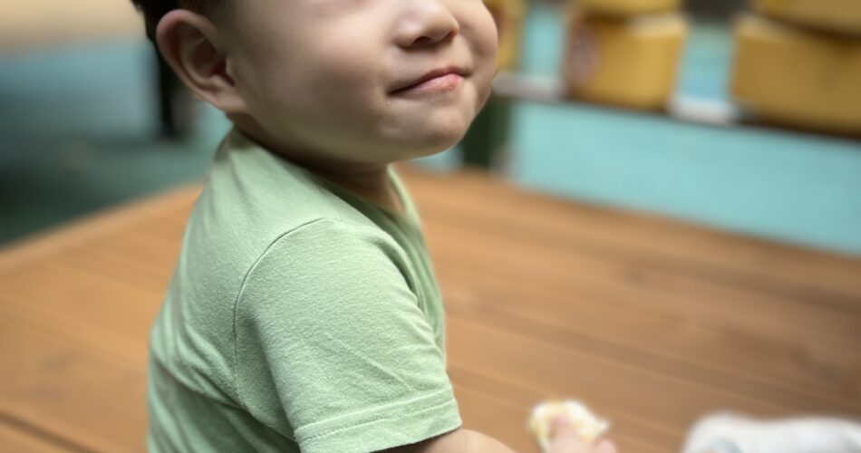 연두색 티셔츠를 입은 아기 지호가 나무 바닥에 앉아 고개를 살짝 들고 오른쪽 위를 바라보며 옅은 미소를 짓고 있다. 배경에는 노란색 블록들로 된 놀이기구가 흐릿하게 보인다.