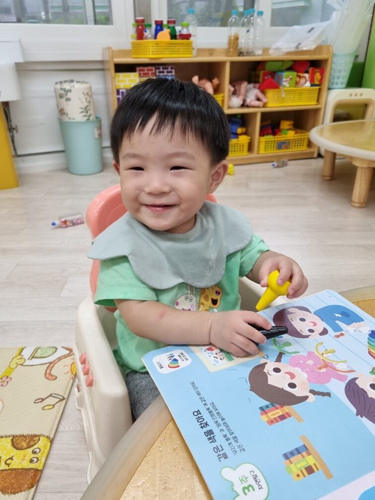 연두색 턱받이와 티셔츠를 입은 지호가 유아용 의자에 앉아 책상 위의 그림책을 보며 펜을 쥐고 환하게 웃고 있다. 배경은 어린이집 교실처럼 보인다.