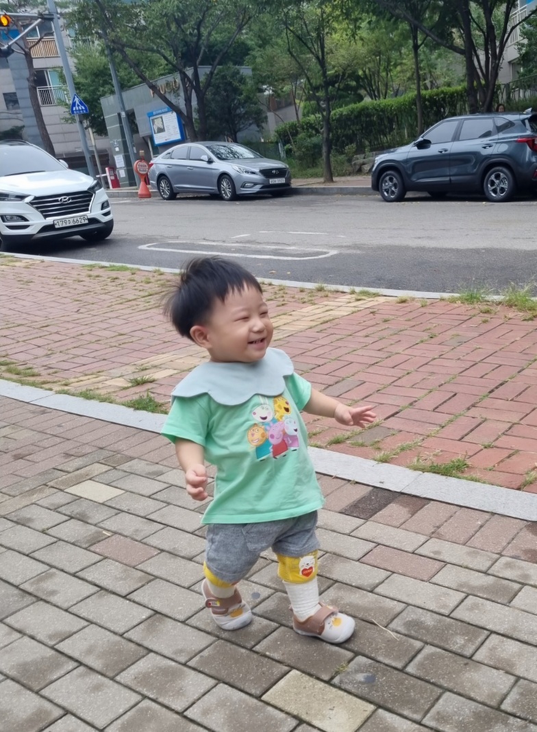 길거리 보도블록 위에서 활짝 웃으며 걷고 있는 지호. 초록색 캐릭터 티셔츠와 회색 반바지, 노란색 무릎 보호대를 착용하고 있다. 뒤로는 주차된 차들이 보인다.