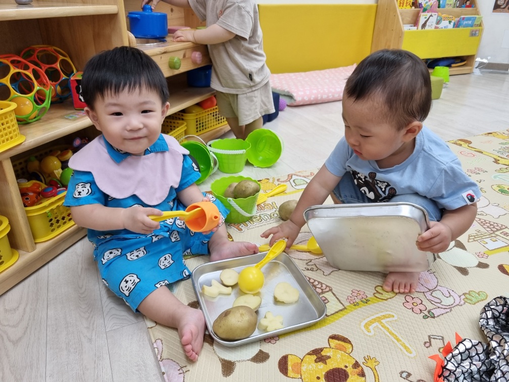 바닥에 앉아 친구와 함께 감자와 여러 도구를 이용해 오감놀이를 하는 지호. 파란색 잠옷과 핑크색 턱받이를 하고 즐거운 표정을 짓고 있다.