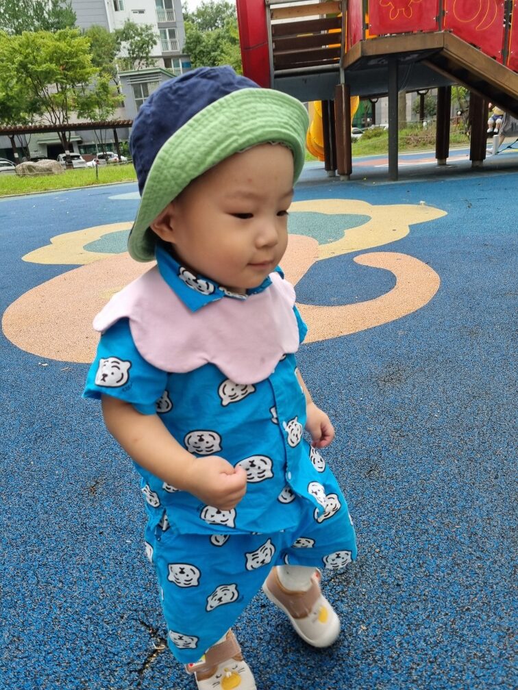 파란색 패턴의 상하의와 모자를 쓴 지호가 파란색 바닥의 놀이터에서 오른쪽으로 걸어가고 있는 모습입니다.