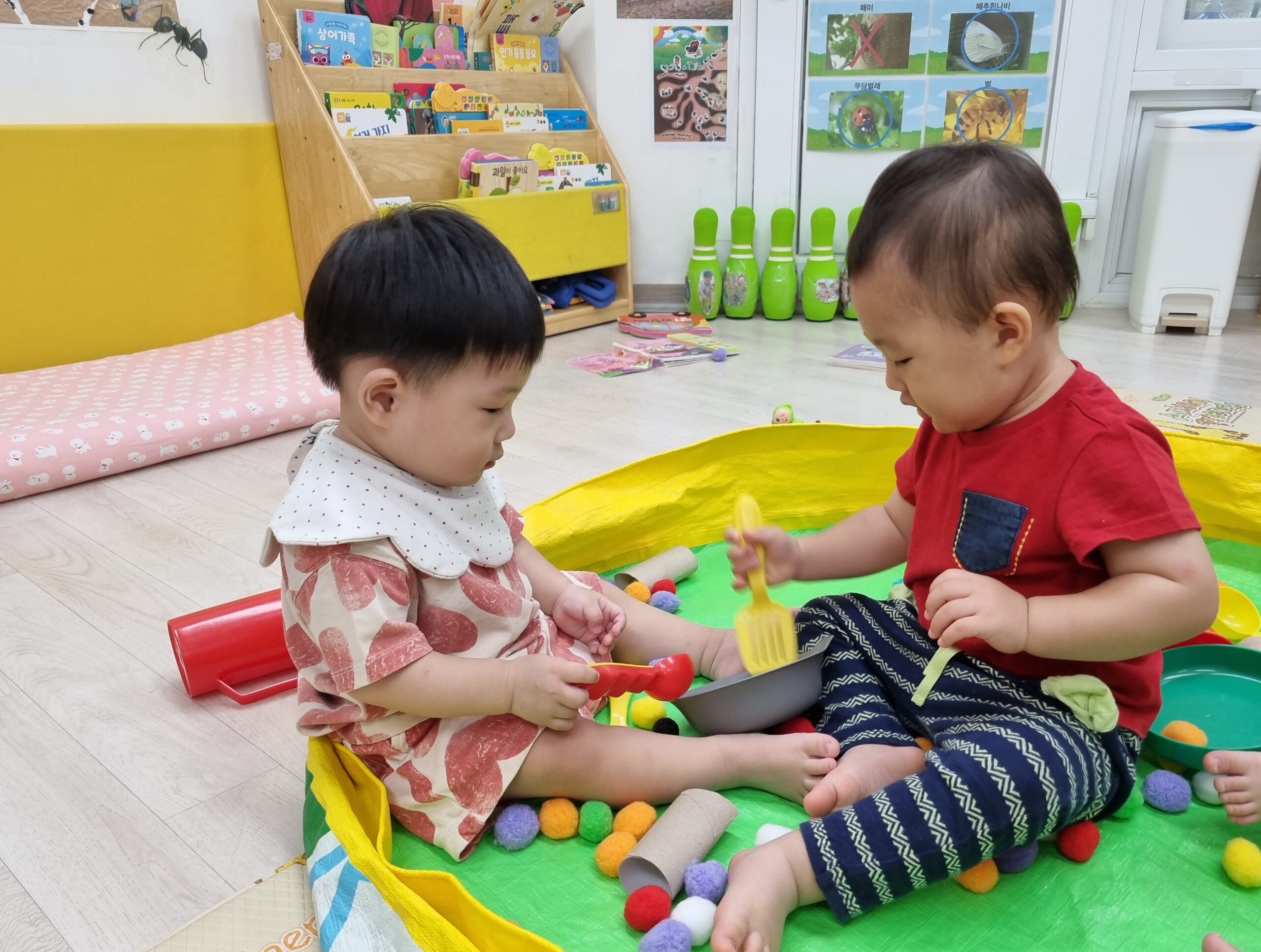 하트 무늬 옷을 입은 지호와 빨간색 옷을 입은 친구가 초록색 매트 위에 앉아 알록달록한 폼폼이들과 작은 그릇, 숟가락으로 서로 마주 보며 놀고 있다. 뒤쪽에는 책꽂이가 보인다.