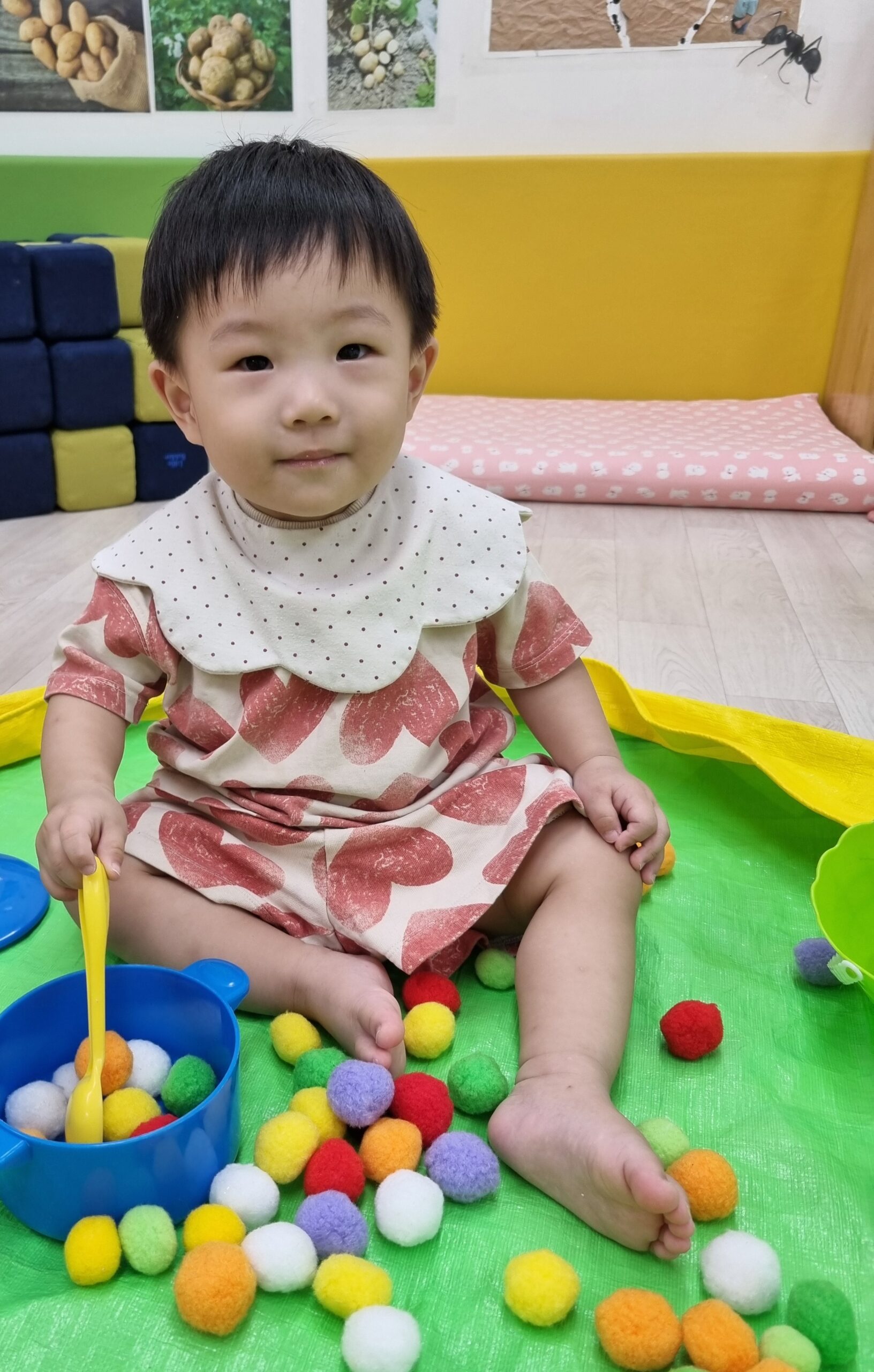 하트 무늬 옷을 입은 지호가 초록색 매트 위 알록달록한 폼폼이들 사이에서 파란색 그릇과 노란색 숟가락을 들고 카메라를 향해 환하게 웃고 있다. 뒤쪽으로는 책꽂이와 노란색 벽이 보인다.