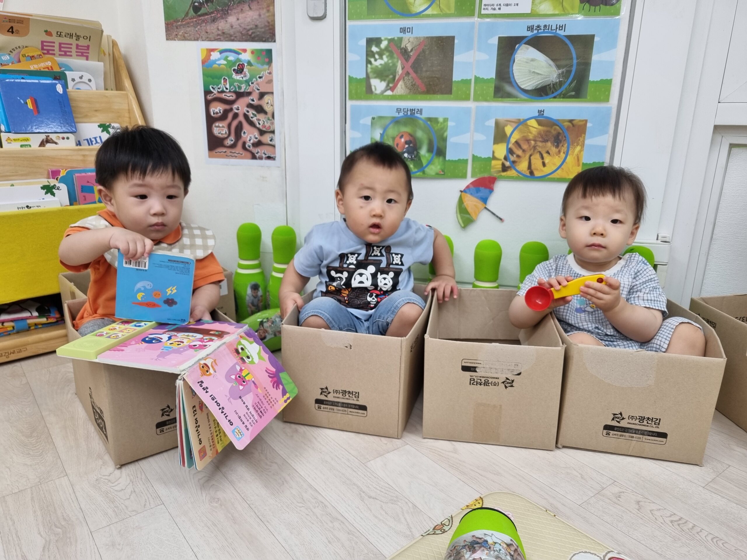 세 명의 아기가 각각 종이 상자 안에 앉아 놀고 있습니다. 왼쪽의 지호는 책을 보고 있고, 가운데 아기는 정면을 응시하며, 오른쪽 아기는 노란색 스푼 장난감을 들고 있습니다. 배경에는 곤충 포스터가 보입니다.