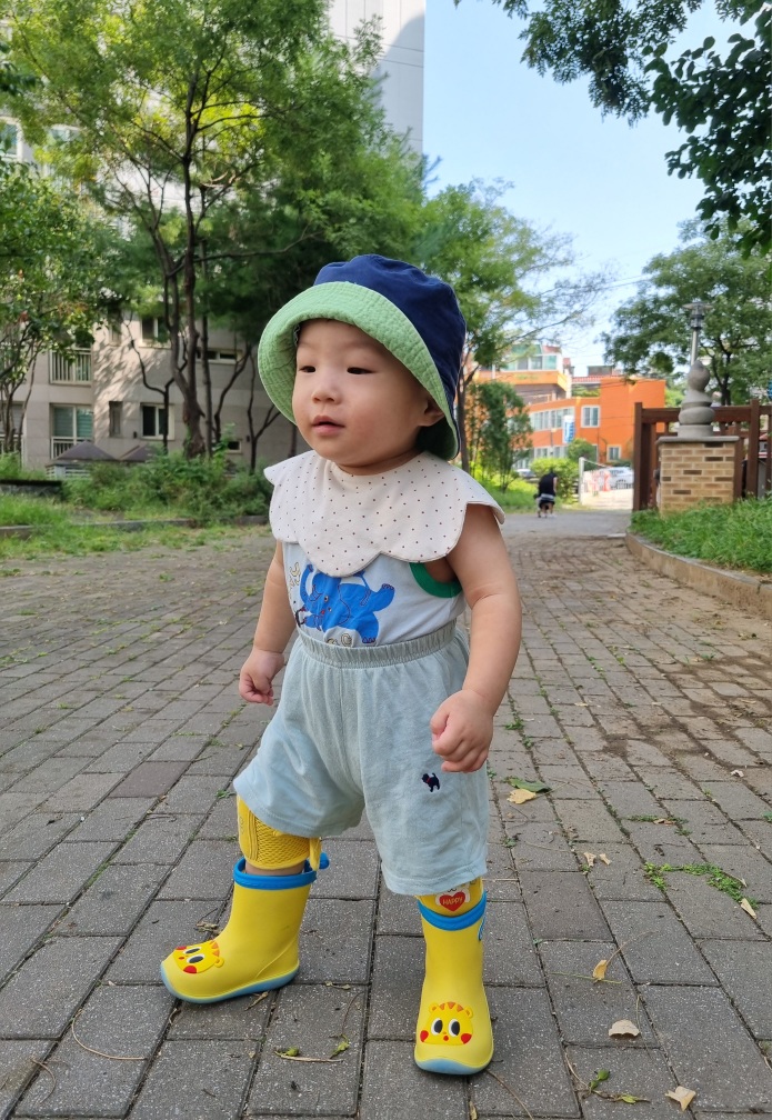 파란색과 초록색이 섞인 모자와 회색 상하의, 그리고 노란색 오리 장화를 신은 지호가 야외 포장도로에 서서 왼쪽으로 시선을 두고 있는 모습입니다.