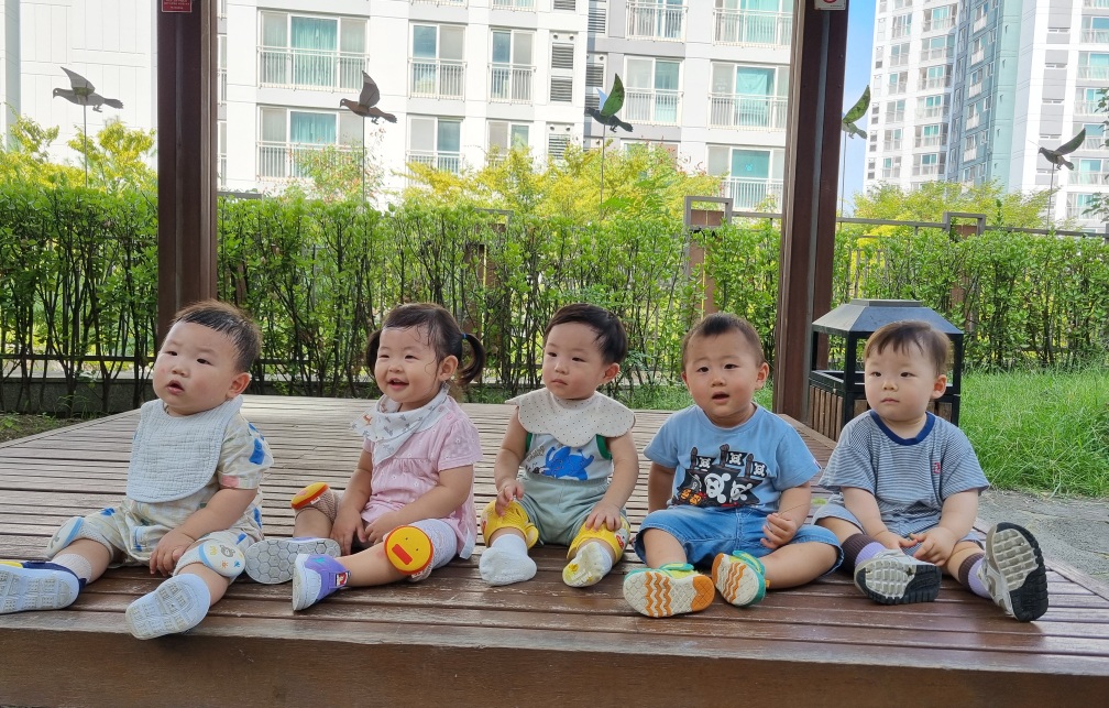 다섯 명의 아기들이 야외 나무 데크에 나란히 앉아있는 모습입니다. 지호는 왼쪽에서 두 번째에 앉아 밝게 웃고 있으며, 배경에는 푸른 덤불과 아파트 건물이 보입니다.
