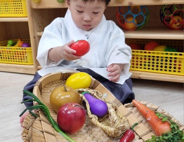 한복을 입은 지호가 바구니에 담긴 가짜 채소들을 탐색하며 빨간 토마토를 손에 쥐고 있는 모습입니다. 배경에는 나무 선반과 장난감들이 보입니다.
