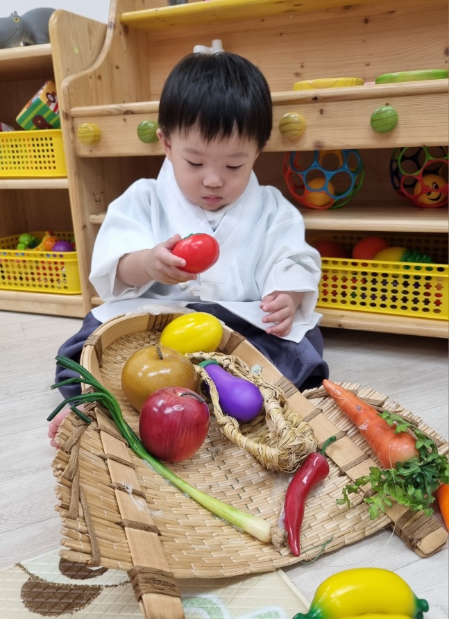 한복을 입은 지호가 바구니에 담긴 가짜 채소들을 탐색하며 빨간 토마토를 손에 쥐고 있는 모습입니다. 배경에는 나무 선반과 장난감들이 보입니다.